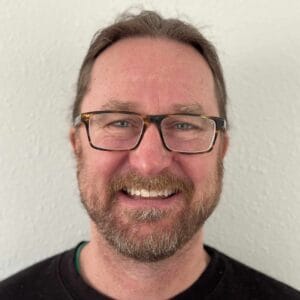 A man with glasses smiling in front of a white wall during the MC Fireside Chats on July 19th, 2023.