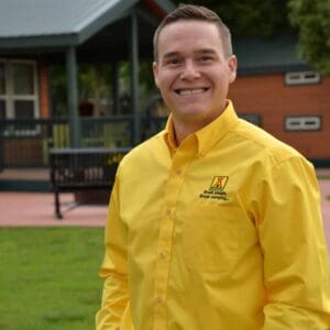 A man in a yellow shirt standing in front of a cabin, delivering MC Fireside Chats on November 15th, 2023.