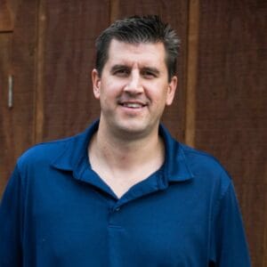 A man in a blue shirt standing in front of a wooden wall during the MC Fireside Chats on November 8th, 2023.