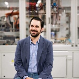 Professional smiling in front of industrial automation equipment during the MC Fireside Chats on March 27th, 2024.