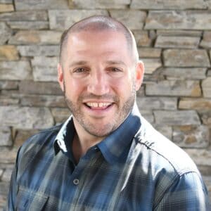 A man in a plaid shirt smiling in front of a stone wall during the MC Fireside Chats on March 6th, 2024.