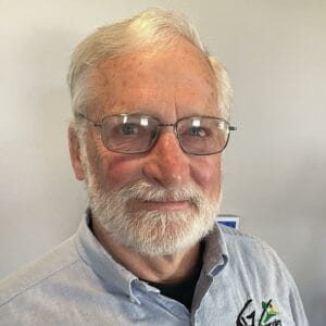 An elderly man with white hair and a beard, wearing glasses and a blue button-up shirt, smiles gently while standing against a plain wall on April 17th, 2024.