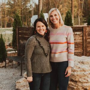 Two smiling women standing together outdoors, one in a green patterned sweater and the other in a striped sweater, with a wooded area in the background during the MC Fireside Chats event.