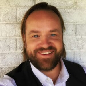A smiling man with a beard, wearing a white shirt and black vest, standing in front of a gray brick wall during the MC Fireside Chats on May 8th, 2024.